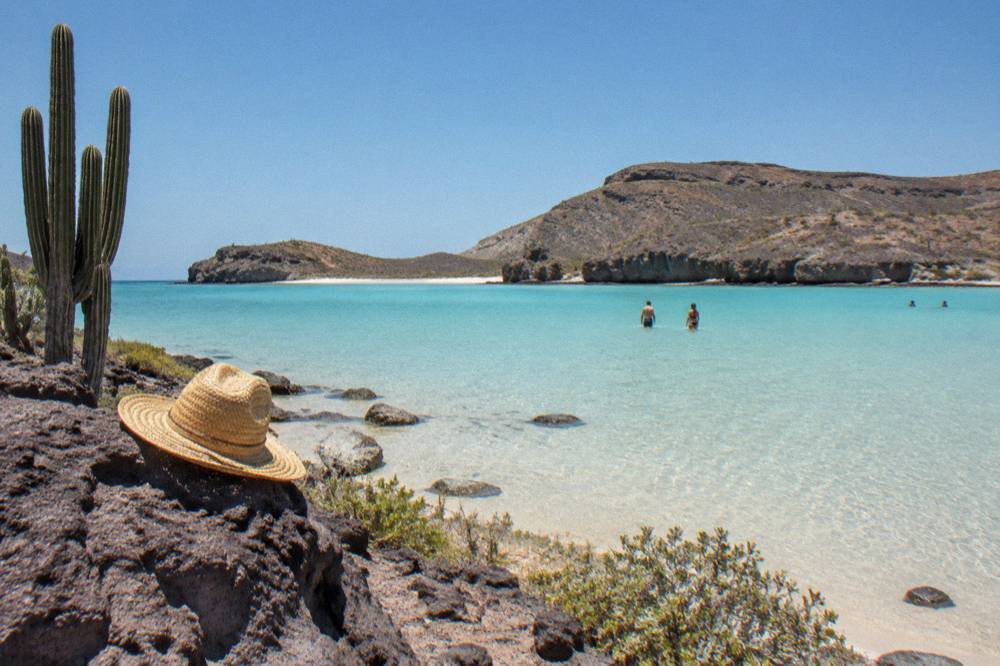 A wide shallow beach of pale sand and turquoise water enclosed by low desert hills