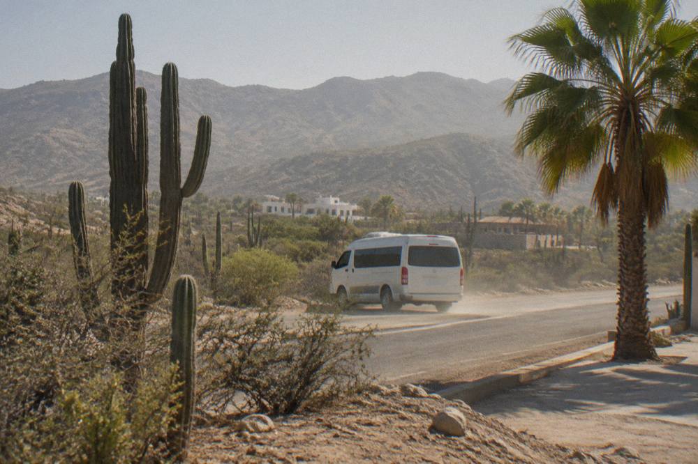 A passenger van traveling a desert highway between cactus-covered hills on the Baja peninsula