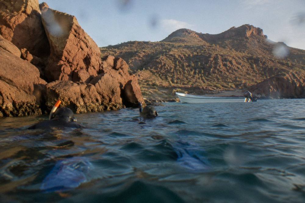 Snorkelers in the water beside a rocky shoreline with a small boat waiting nearby