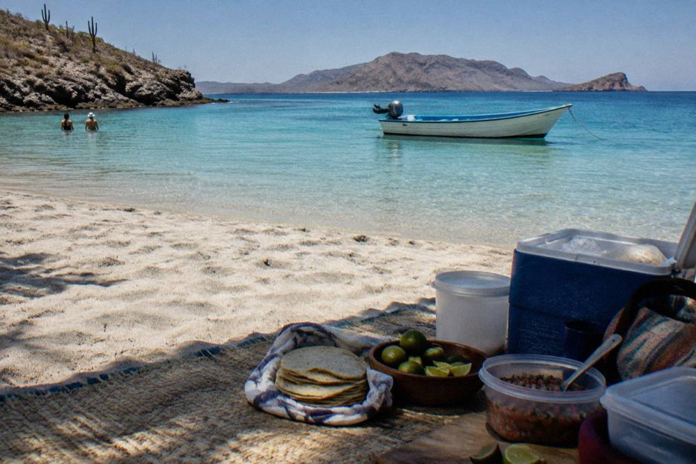 A picnic lunch laid out on a beach with a boat anchored in clear shallow water