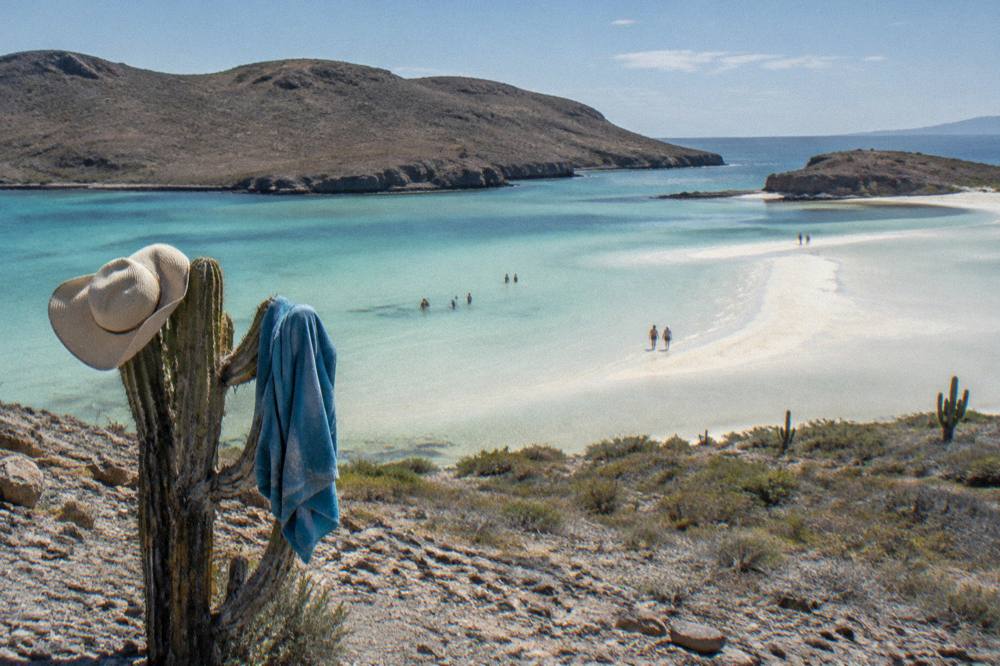 A shallow turquoise bay ringed by low hills with a sandy spit at low tide