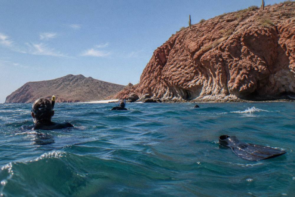 Snorkelers in the water beside tall rocky cliffs at an island in blue open sea
